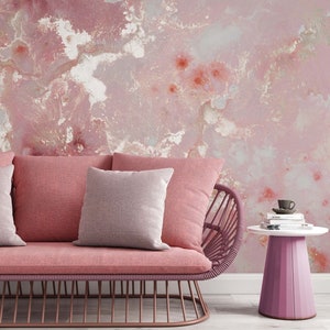 May include: A pink wicker sofa with two pillows, one pink and one white, sits in front of a pink and white marbled wall. A small pink side table with a stack of books and a cup on top sits to the right of the sofa.