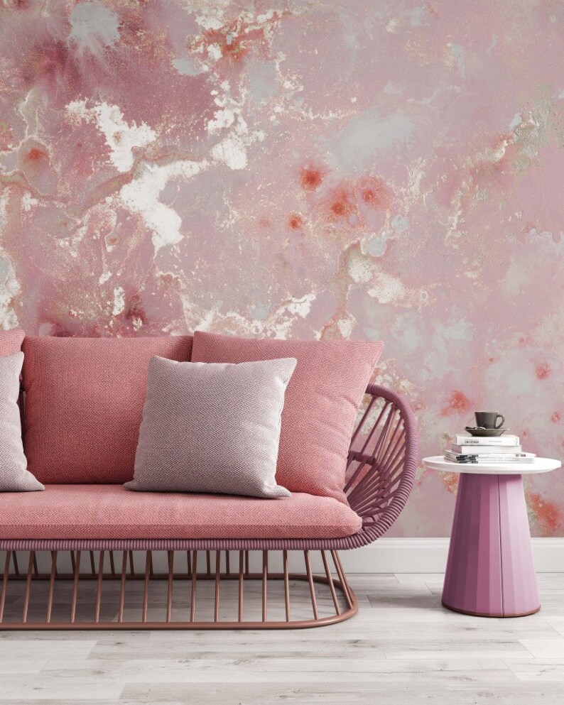 May include: A pink wicker sofa with two pillows, one pink and one white, sits in front of a pink and white marbled wall. A small pink side table with a stack of books and a cup on top sits to the right of the sofa.