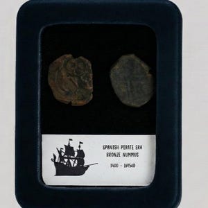 May include: A dark blue display case holds two antique bronze coins. The case features a label with a silhouette of a sailing ship and the text "SPANISH PIRATE ERA BRONZE NUMMUS 1400 - 1695AD". The coins are brown and have visible details.