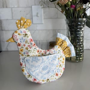 K&ouml;nnte beinhalten: Ein k&uuml;chenaccessoire in Form eines Stoffhuhns mit einem Blumenmuster in Rosa, Gelb und Blau. Das Huhn hat einen gelben Kamm und Schnabel sowie eine blaue Blumentasche. Im Hintergrund steht eine Vase mit Blumen.