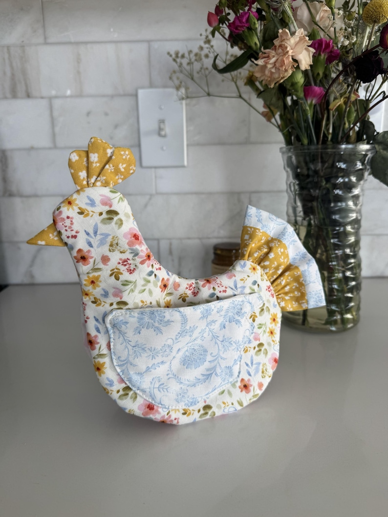 K&ouml;nnte beinhalten: Ein k&uuml;chenaccessoire in Form eines Stoffhuhns mit einem Blumenmuster in Rosa, Gelb und Blau. Das Huhn hat einen gelben Kamm und Schnabel sowie eine blaue Blumentasche. Im Hintergrund steht eine Vase mit Blumen.