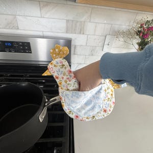 K&ouml;nnte beinhalten: Ein huhnf&ouml;rmiger Ofenhandschuh mit floralem Muster, gelbem Kamm und Schnabel. Der Handschuh hat ein blau-wei&szlig;es Blumenmuster auf der Vorderseite und ein gelb-wei&szlig;es Blumenmuster auf der R&uuml;ckseite. Der Handschuh wird in der N&auml;he eines schwarzen Topfes auf einem Herd gehalten.