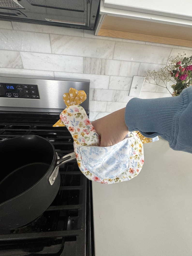 K&ouml;nnte beinhalten: Ein huhnf&ouml;rmiger Ofenhandschuh mit floralem Muster, gelbem Kamm und Schnabel. Der Handschuh hat ein blau-wei&szlig;es Blumenmuster auf der Vorderseite und ein gelb-wei&szlig;es Blumenmuster auf der R&uuml;ckseite. Der Handschuh wird in der N&auml;he eines schwarzen Topfes auf einem Herd gehalten.