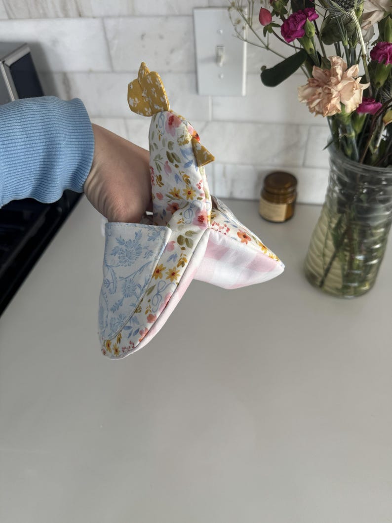 K&ouml;nnte beinhalten: Ein handgemachter Ofenhandschuh in Form eines Huhns. Der Handschuh hat einen gelben Kamm und Stoff mit Blumenmuster in Wei&szlig;, Gelb und Rosa. Die Innenseite ist mit einem rosa-wei&szlig; gestreiften Stoff gef&uuml;ttert, und die Manschette hat ein blaues Blumenmuster.