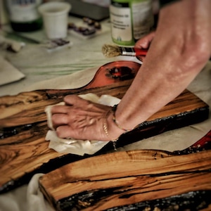 Può includere: Una persona applica una finitura a un tagliere di legno. La tavola è rettangolare con un bordo vivo e una resina epossidica tinta di rosso. La venatura del legno è visibile e la tavola viene pulita con un panno bianco. L'immagine è leggermente sfocata.