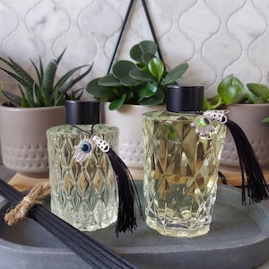 May include: Two clear glass diffuser bottles with black lids and decorative charms. The bottles have a textured diamond pattern and are filled with a light yellow liquid. The bottles are sitting on a grey oval tray with black diffuser sticks.