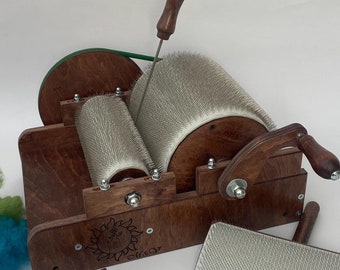 Könnte beinhalten: Ein Trommelkardiergerät aus Holz mit zwei metallborstigen Walzen, einer Handkurbel und einem vertikalen Griff. Das Gerät verfügt über ein Sonnenlogo, eine separate rechteckige Handkarde, eine Metallklemme und kleine Büschel aus gelber, grüner und blauer Rohwolle.