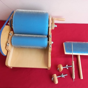 Puede incluir: Una m&aacute;quina de cardar lana de madera con dos rodillos azules y un mango de madera. La m&aacute;quina est&aacute; sobre una superficie roja con un peine de cardar azul y dos pomos de madera con tornillos de metal.