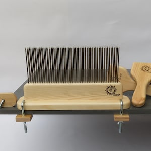 May include: A wooden wool carding set with a metal comb and two wooden carding tools. The tools are clamped to a wooden surface. The tools have a sun design with the text "Carding" on them.