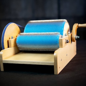 Puede incluir: Un cardador de lana de madera con un rodillo azul y blanco y un mango de madera. El cardador se utiliza para limpiar y preparar la lana para hilar.