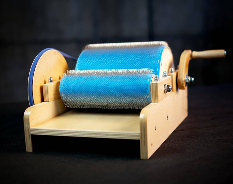 Puede incluir: Un cardador de lana de madera con un rodillo azul y blanco y un mango de madera. El cardador se utiliza para limpiar y preparar la lana para hilar.