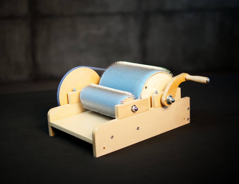 Puede incluir: Un torno de hilar de madera con un rodillo de cepillo azul y un mango de madera. El torno est&aacute; sobre una superficie negra.