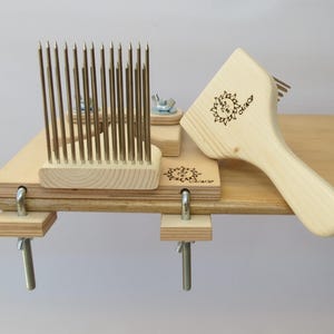 Op de afbeelding: Een houten wollen kaardstel met twee kaardgereedschappen en een houten borstel. De kaardgereedschappen hebben rijen metalen tanden en zijn met klemmen aan een houten basis bevestigd. De borstel heeft een houten handvat en een rechthoekige borstelkop.