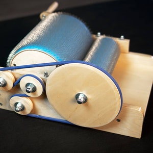 Puede incluir: Un torno de hilar de madera con una correa de goma azul y un rodillo de aguja de metal. El torno est&aacute; hecho de madera clara y tiene un acabado natural.