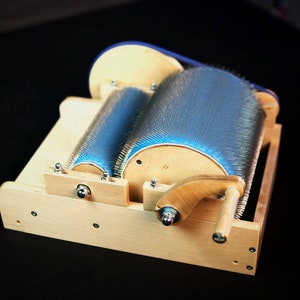 Puede incluir: Un cardador de lana de madera con dos rodillos de cerdas azules. El cardador tiene un mango de madera y est&aacute; hecho de madera clara.