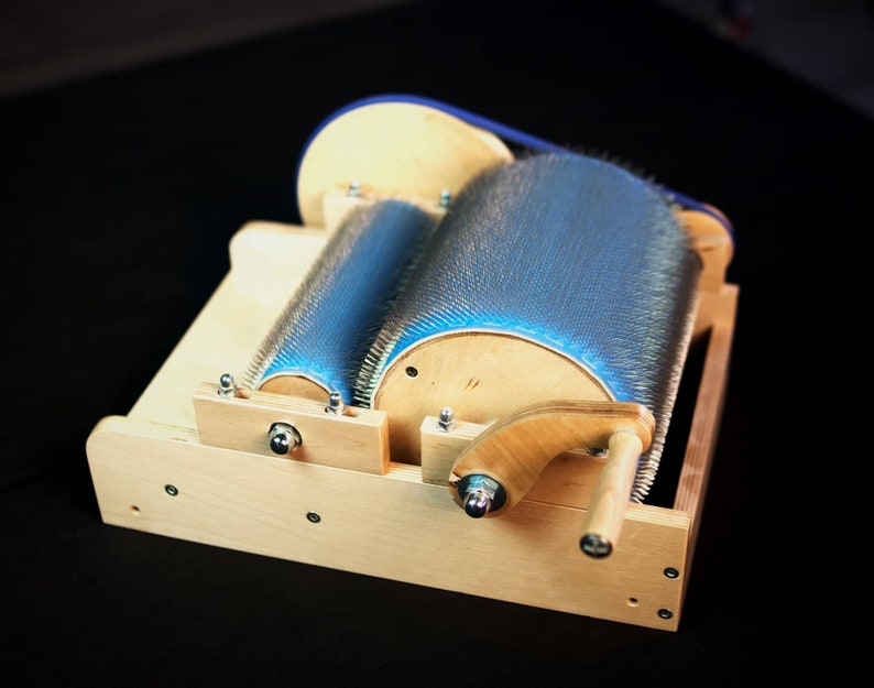 Puede incluir: Un cardador de lana de madera con dos rodillos de cerdas azules. El cardador tiene un mango de madera y est&aacute; hecho de madera clara.