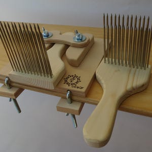 May include: A wooden fiber comb with two comb heads and a wooden base. The comb heads have multiple metal tines. The base has a sun logo and the word "Olgaq" etched into the wood. The comb is designed for fiber preparation.