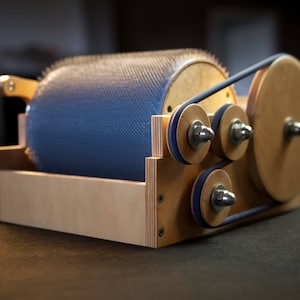 Puede incluir: Un torno de hilar de madera con un tambor azul y un sistema de transmisi&oacute;n por correa. El tambor tiene una superficie texturizada y est&aacute; cubierto de peque&ntilde;as cerdas blancas. El torno est&aacute; hecho de madera clara y tiene un acabado natural.