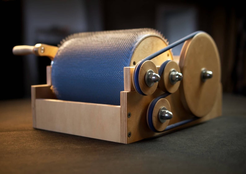 Puede incluir: Un torno de hilar de madera con un tambor azul y un sistema de transmisi&oacute;n por correa. El tambor tiene una superficie texturizada y est&aacute; cubierto de peque&ntilde;as cerdas blancas. El torno est&aacute; hecho de madera clara y tiene un acabado natural.