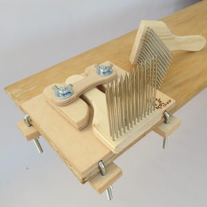 May include: A wooden wool carding tool with metal teeth. The carding tool is attached to a wooden base with clamps. The tool is used to blend and prepare wool for spinning.
