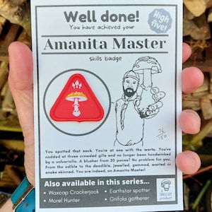 May include: A white card with the text "Well done! You have achieved your Amanita Master skills badge" and a red triangle patch with a white mushroom illustration. The card also shows a cartoon illustration of a person holding a mushroom. The text "Also available in this series..." is listed with bullet points: "Waxcap Crackerjack", "Earthstar spotter", "Morel Hunter", and "Grifola gatherer".
