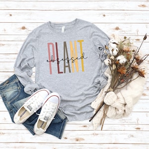 May include: A gray long-sleeve shirt with the text "PLANT obsessed" in colorful letters. The shirt is laid out on a white wood background with a pair of white sneakers and a bouquet of dried flowers.