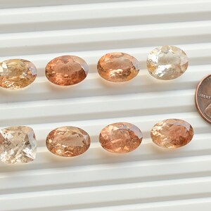 May include: Ten loose oval and square cut gemstones in shades of peach, pink, and brown. The stones are arranged in two rows on a white surface. A US penny is included for scale.