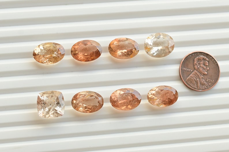 May include: Ten loose oval and square cut gemstones in shades of peach, pink, and brown. The stones are arranged in two rows on a white surface. A US penny is included for scale.