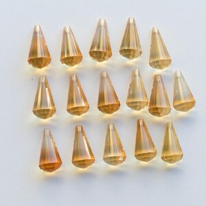 Puede incluir: Una colecci&oacute;n de 18 cuentas de cristal de color &aacute;mbar en forma de l&aacute;grima con un dise&ntilde;o facetado. Las cuentas est&aacute;n dispuestas en filas sobre una superficie blanca.