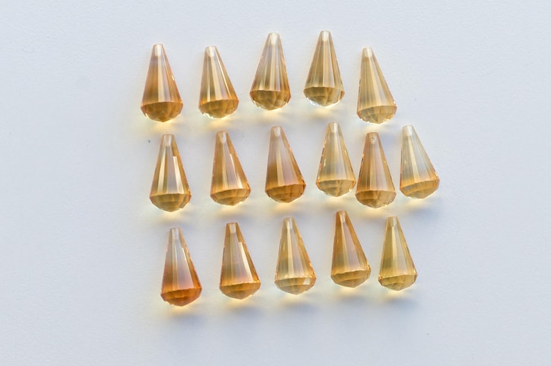 Puede incluir: Una colecci&oacute;n de 18 cuentas de cristal de color &aacute;mbar en forma de l&aacute;grima con un dise&ntilde;o facetado. Las cuentas est&aacute;n dispuestas en filas sobre una superficie blanca.