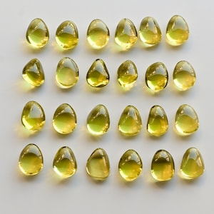 May include: A collection of twenty-four yellow, teardrop-shaped capsules arranged in rows on a white surface. The capsules have a translucent appearance, with a slight sheen, and are evenly spaced.