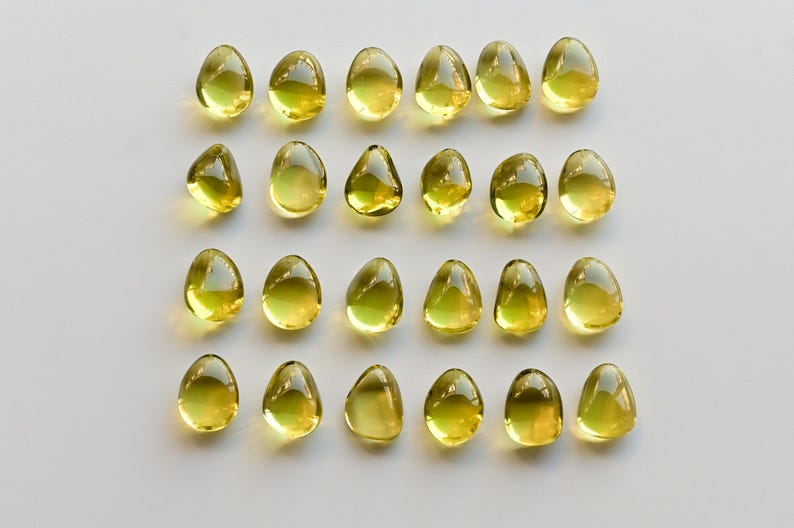 May include: A collection of twenty-four yellow, teardrop-shaped capsules arranged in rows on a white surface. The capsules have a translucent appearance, with a slight sheen, and are evenly spaced.