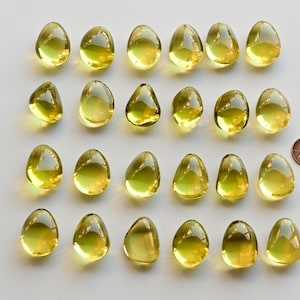 May include: Twenty-four translucent, yellow-green, teardrop-shaped glass beads are arranged on a white surface. A copper penny is next to the beads for size comparison. The beads have a smooth, glossy finish.