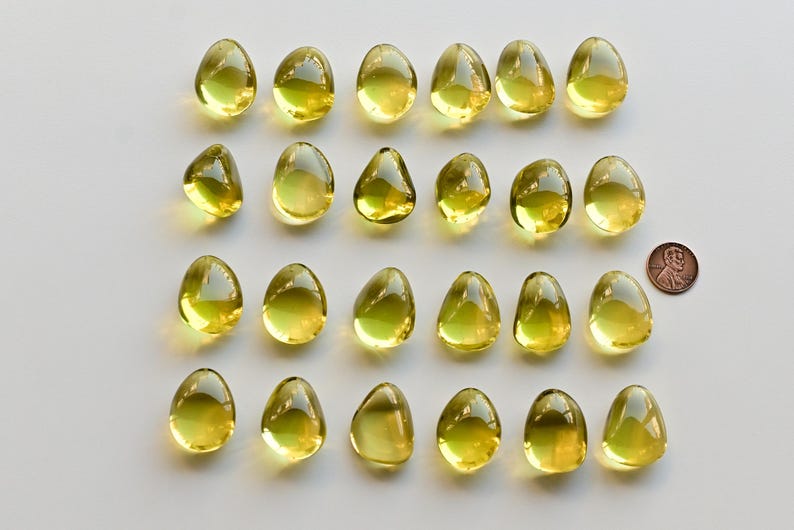 May include: Twenty-four translucent, yellow-green, teardrop-shaped glass beads are arranged on a white surface. A copper penny is next to the beads for size comparison. The beads have a smooth, glossy finish.
