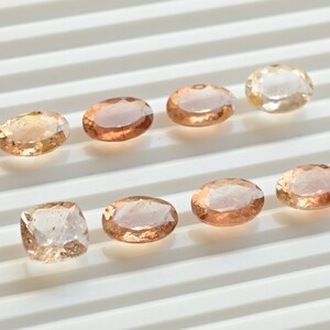 May include: Assortment of eight oval-shaped and one square-shaped gemstones in shades of light brown and peach. The gemstones are arranged in two rows on a white surface.