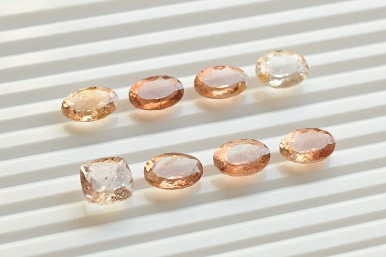 May include: Assortment of eight oval-shaped and one square-shaped gemstones in shades of light brown and peach. The gemstones are arranged in two rows on a white surface.