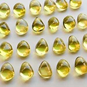 May include: A collection of translucent, teardrop-shaped objects with a yellow-green hue. The objects are arranged on a white surface, reflecting light and creating highlights. The overall aesthetic is clean and minimalist.