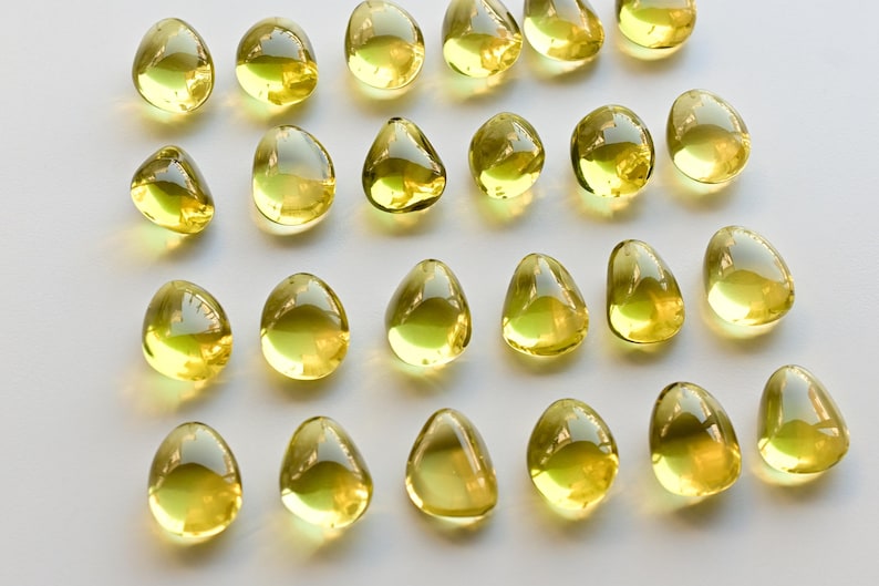 May include: A collection of translucent, teardrop-shaped objects with a yellow-green hue. The objects are arranged on a white surface, reflecting light and creating highlights. The overall aesthetic is clean and minimalist.