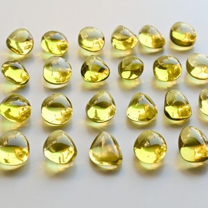 May include: A collection of twenty-four yellow, teardrop-shaped glass beads. The beads are arranged in a grid pattern on a white surface. Each bead has a smooth, rounded form and a glossy finish, reflecting light.