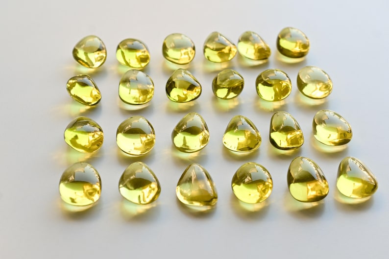 May include: A collection of twenty-four yellow, teardrop-shaped glass beads. The beads are arranged in a grid pattern on a white surface. Each bead has a smooth, rounded form and a glossy finish, reflecting light.