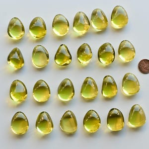 May include: Twenty-one translucent, teardrop-shaped, yellow-green glass beads are arranged on a white surface. A copper penny is next to the beads for size comparison. The beads have a smooth, glossy finish and vary slightly in size and shape.