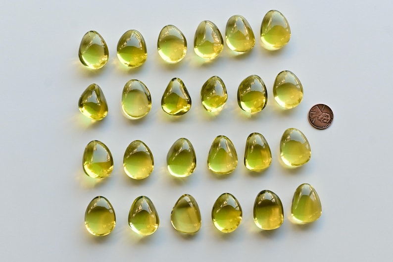 May include: Twenty-one translucent, teardrop-shaped, yellow-green glass beads are arranged on a white surface. A copper penny is next to the beads for size comparison. The beads have a smooth, glossy finish and vary slightly in size and shape.