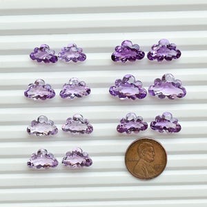 May include: A collection of amethyst cloud-shaped gemstones. The gemstones are a translucent purple color and are arranged in rows on a white, ridged surface. A penny is included for size reference.