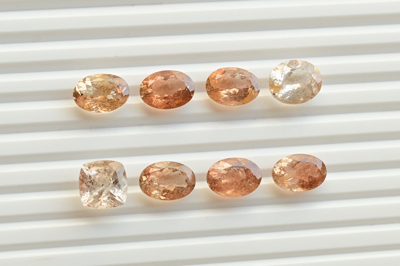 May include: Eight oval-shaped and one square-shaped gemstones in shades of brown and pink. The gemstones are arranged in two rows on a white surface.
