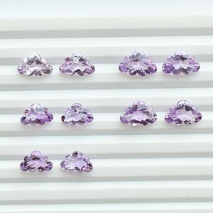 May include: A selection of cloud-shaped amethyst gemstones. The gemstones are a pale purple hue and are arranged in rows on a white, ridged surface. The facets of the stones reflect light, creating a sparkling effect. These are suitable for jewellery making.