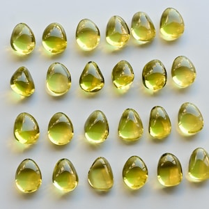 May include: A collection of twenty-one translucent, teardrop-shaped, yellow-green gemstones arranged on a white surface. Each gem has a smooth, polished appearance, reflecting light. The gems vary slightly in size and shape, creating a visually appealing pattern.