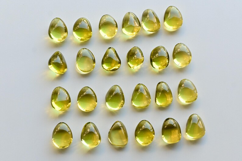 May include: A collection of twenty-one translucent, teardrop-shaped, yellow-green gemstones arranged on a white surface. Each gem has a smooth, polished appearance, reflecting light. The gems vary slightly in size and shape, creating a visually appealing pattern.