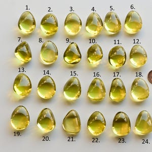 May include: A collection of twenty-four yellow-green, teardrop-shaped gemstones, numbered 1 through 24, are displayed on a white surface. A penny is included for size comparison. The gemstones vary slightly in shape and clarity.