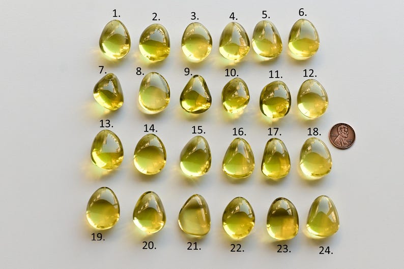 May include: A collection of twenty-four yellow-green, teardrop-shaped gemstones, numbered 1 through 24, are displayed on a white surface. A penny is included for size comparison. The gemstones vary slightly in shape and clarity.