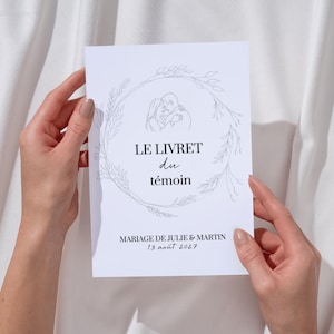 Peut inclure: Un livret de mariage blanc avec le texte "LE LIVRET du témoin" et "MARIAGE DE JULIE & MARTIN 13 août 2027". Le livret présente un dessin au trait d'un couple s'embrassant dans une couronne florale.